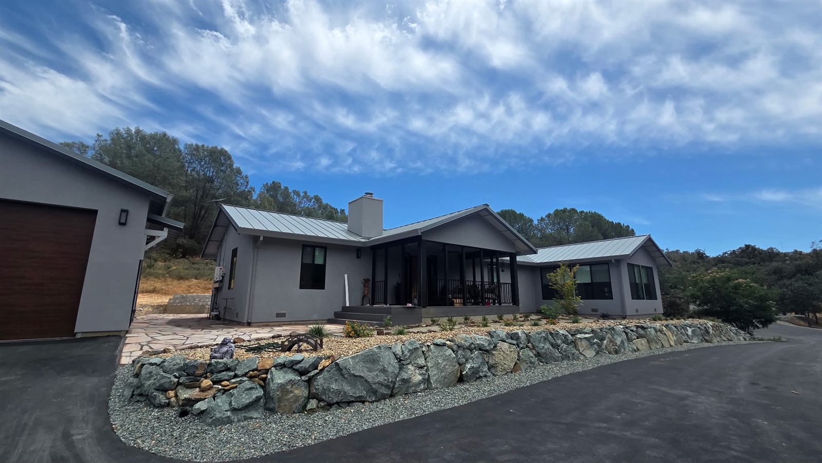 Grey ranch-style home with a standing-seam metal roof behind a new green-grey boulder retaining wall, gravel beds, young plantings and a flagstone approach
