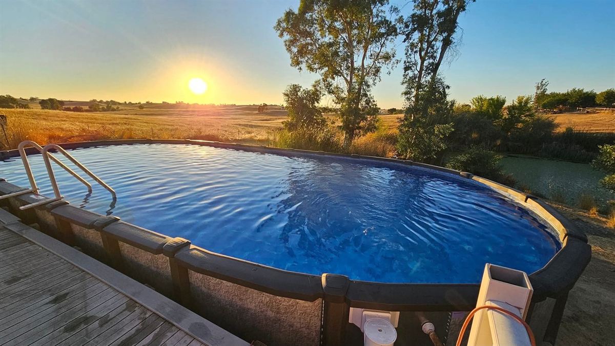 Large oval above-ground pool with a stone-look resin frame and metal entry ladder, set beside a timber deck, filled and still, with golden grassland and a sunset behind