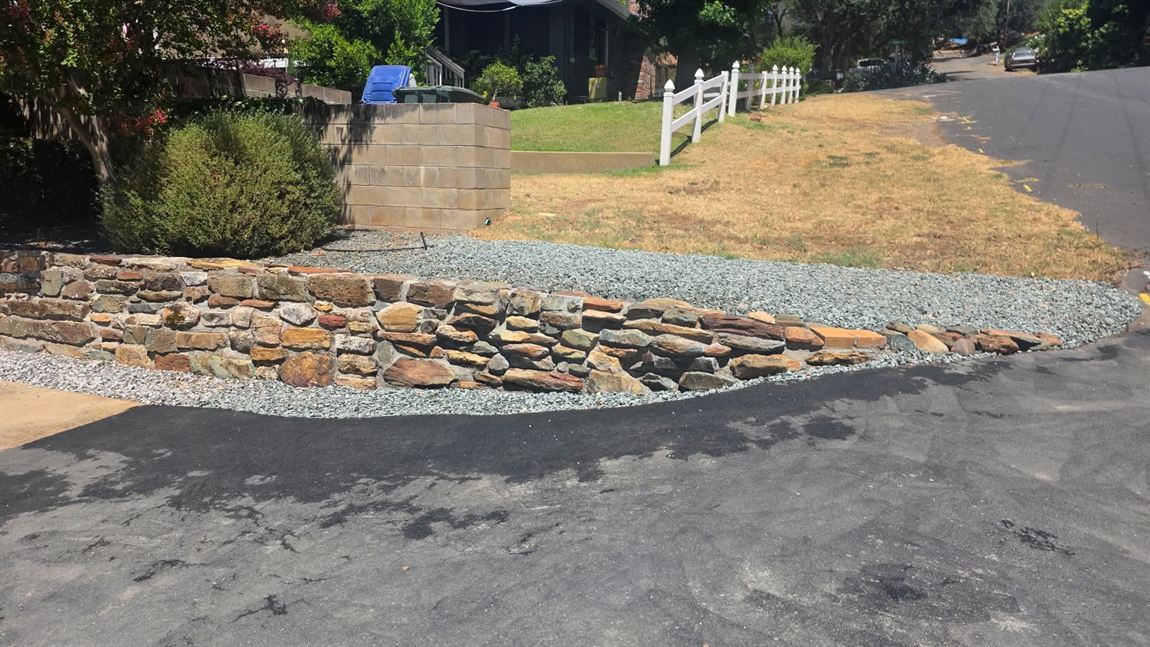 Long low dry-stacked natural stone wall in gold and rust Sierra fieldstone curving along the edge of an asphalt driveway, backed by a bed of grey-green crushed rock