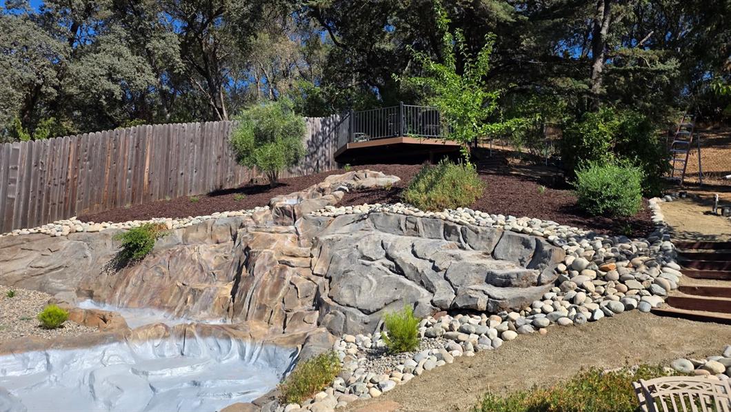 Sculpted multi-tier rock waterfall and stream channel built into a slope, edged with rounded river cobble and bark-mulched planting beds below a timber deck
